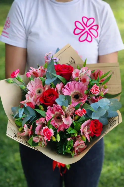 Ramo de flores con gerberas rosas, rosas rojas y claveles rosados, envuelto en papel kraft vintage con diseño impreso, sostenido por una persona frente a un fondo natural de grama verde. Ideal para regalos, floristerías o campañas de publicidad emocional en redes sociales. - 1013050124